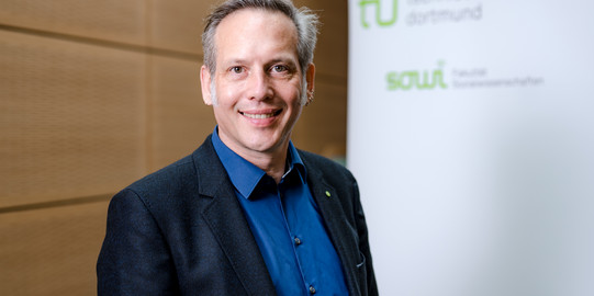 Prof. Dr. Cornelius Schubert Das Foto zeigt das Porträt von Prof. Dr. Cornelius Schubert. Hinter ihm steht ein Rollup auf dem die Logos der Technischen Universität Dormund und der Fakultät Sozialwissenschaften zu lesen sind.